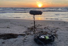 Im Vordergrund sieht man einen Mikrofon Aufbau in einem Blimb als Windschutz auf einem Stativ. Im Hintergrund ist die Ostsee und der beginnende Aufgang der Sonne zu sehen.