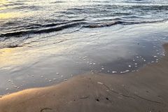 Das Bild zeigt wie kleine Wellen auf den Strand schwappen. Es ist eine Detailaufnahme mit Sand, Wasser und Resten von Seegras auf dem Sand